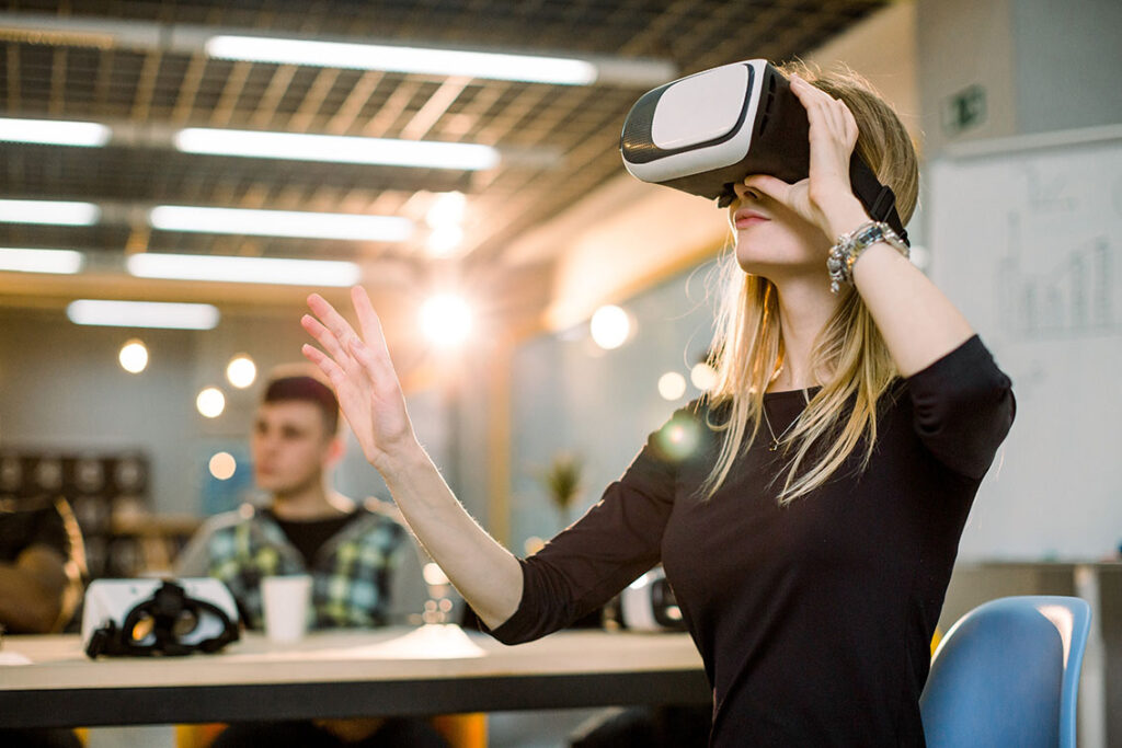 photo d'une femme assise à son bureau. Elle utilise un casque de réalité virtuelle. A l'arrière-plan, un homme est assis à un autre bureau.